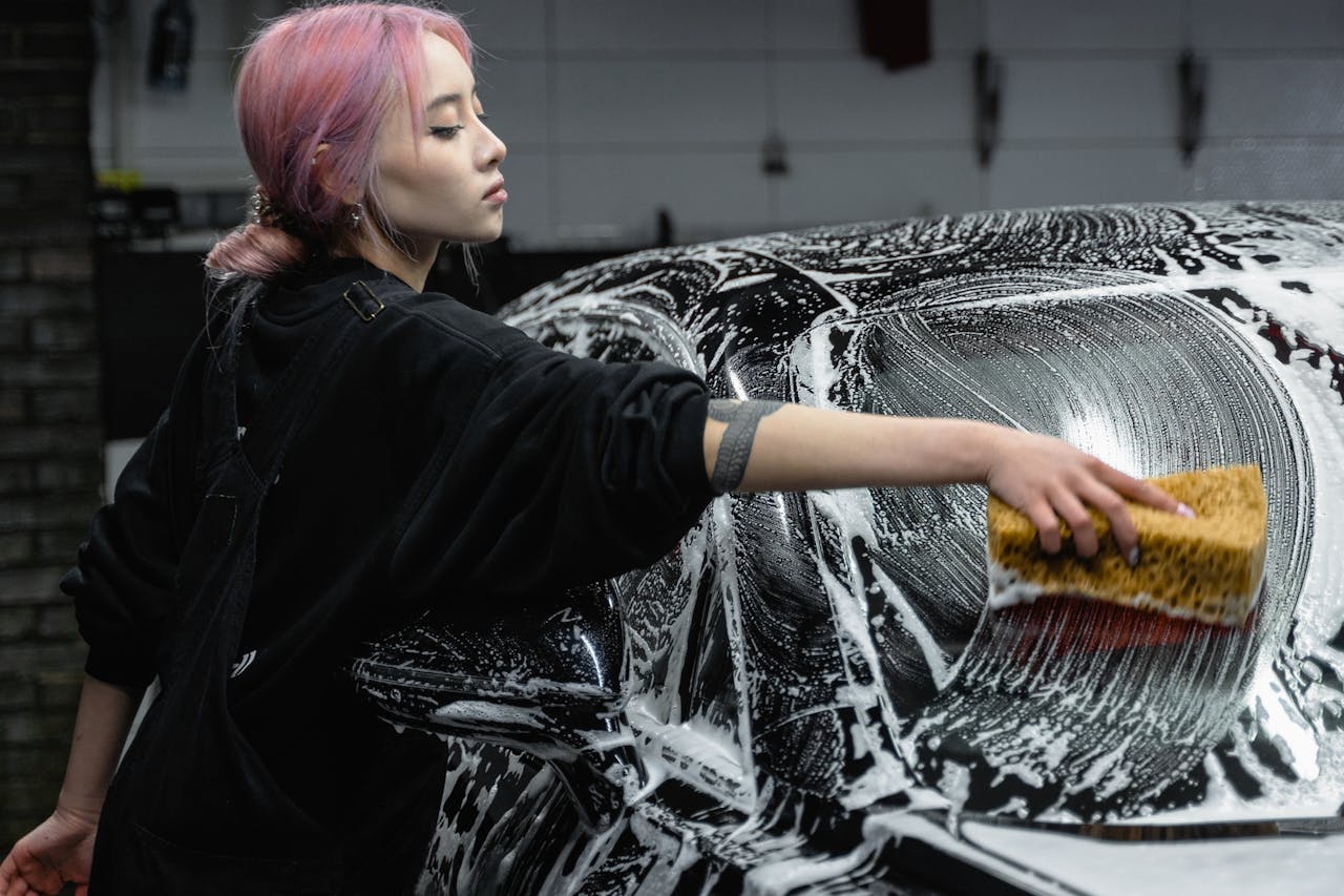 A young woman cleans a black car with a sponge and soap in a garage setting, showing care and attention.